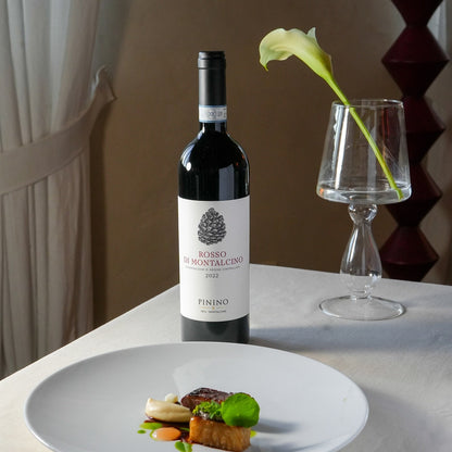 A bottle of Rosso La Montalcino Pinino red wine stands on a white tablecloth next to a stemmed glass with a calla lily stem, with a plated gourmet food dish in the foreground.